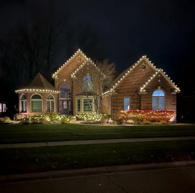 A brick ranch with every peak outlined in warm white, plus lit shrubs and a red-lit hedge.