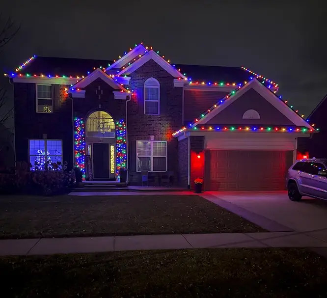 A two-story brick home outlined in multicolor lights, with lit porch columns and a red-lit garage.
