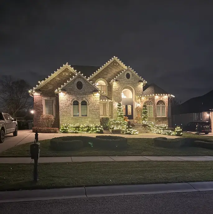 A stone and brick home outlined in warm white lights, with lit evergreens and bushes along the walk.